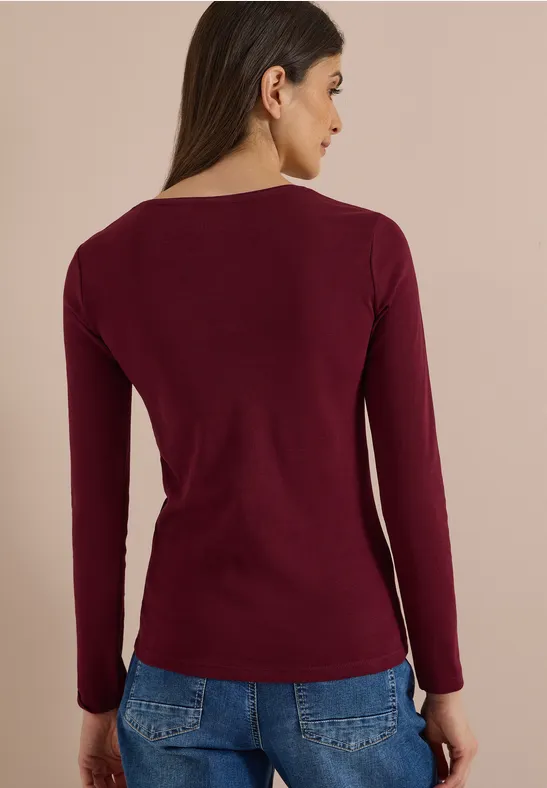Frau von hinten in weinrotem Langarmshirt vor einfarbigem Hintergrund.
