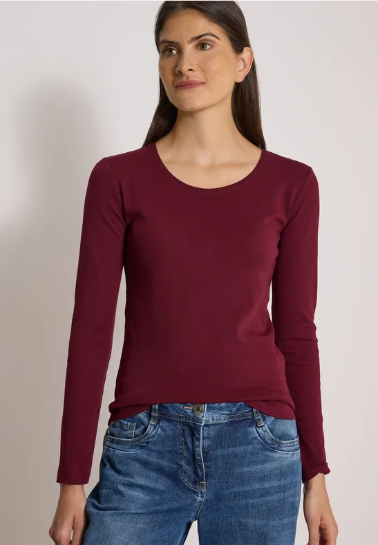 Junge Frau in bordeauxfarbenem Langarmshirt und Jeans vor neutralem Hintergrund.