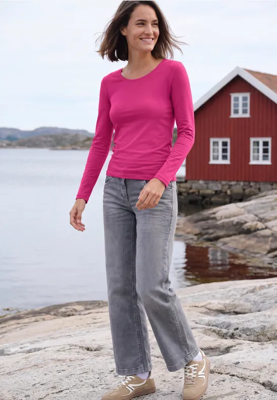 Eine Frau in rosa Pullover und Jeans spaziert lächelnd am Felsufer vor rotem Haus und Meer.