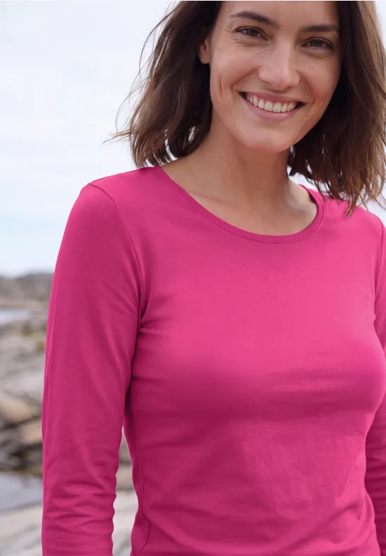 Fröhliche Frau in langärmeligem pinken Shirt vor unscharfem Hintergrund am Meer.