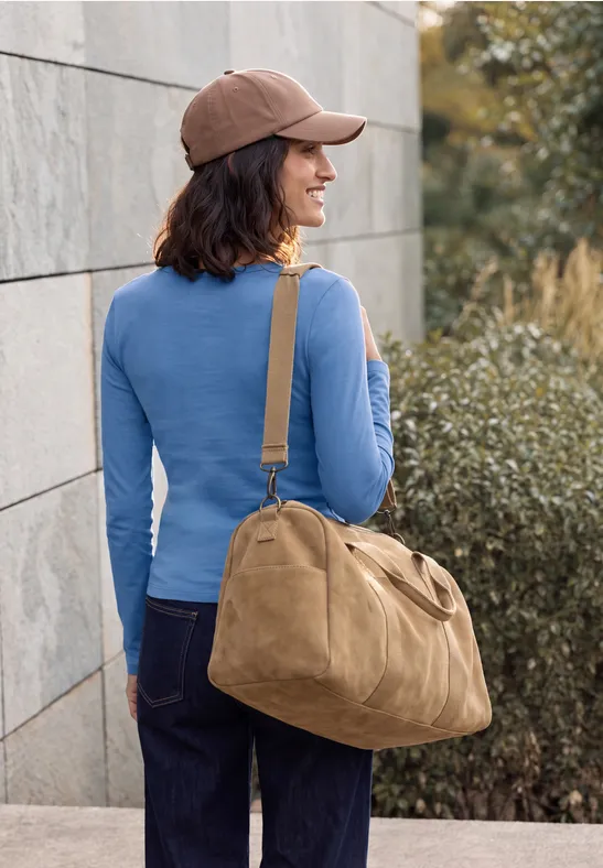 Frau in blauem Oberteil, brauner Kappe und Jeans trägt große beige Tasche und steht vor einer Wand.