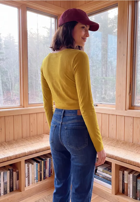 Eine Frau in roter Cap, gelbem Langarmshirt und Jeans blickt über die Schulter in einem hellen Holzraum.