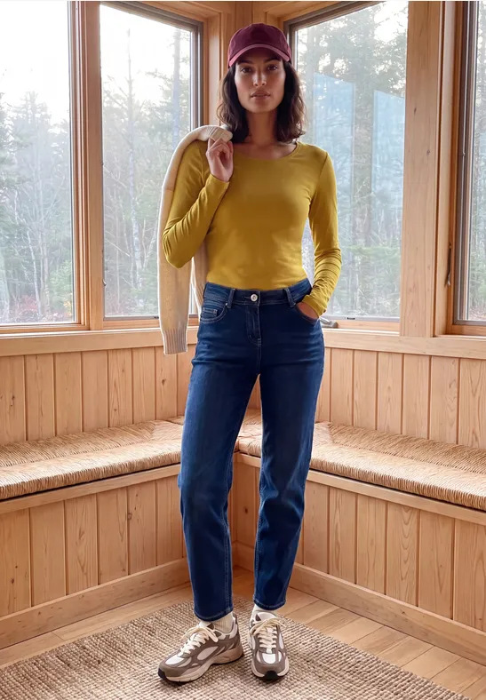 Frau mit gelbem Pullover, blauen Jeans und Mütze steht in einem hellen, holzvertäfelten Raum.