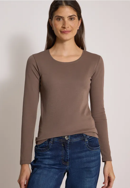 Eine Frau trägt ein braunes Langarmshirt und blaue Jeans und lächelt vor schlichtem Hintergrund.