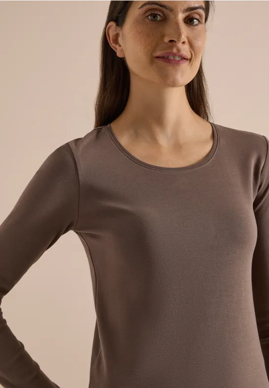 Eine Frau mit braunem Langarmshirt vor beigem Hintergrund, die lächelnd in die Kamera blickt.