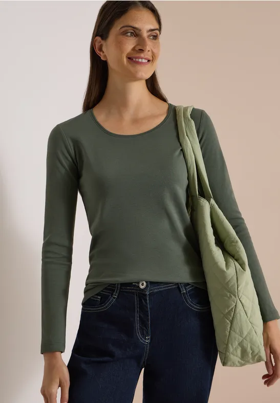 Lächelnde Frau in grünem Langarmshirt und gesteppter Schultertasche vor neutralem Hintergrund.