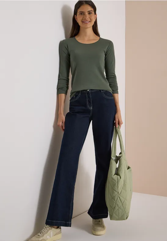 Frau in grünem Langarmshirt, weiten Jeans und beigen Schuhen trägt eine gesteppte grüne Tasche.