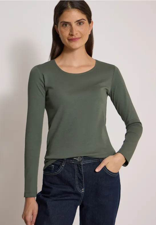 Junge Frau in schlichtem, olivgrünen Langarmshirt und blauen Jeans vor neutralem Hintergrund.