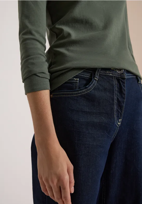 Person in dunkelgrünem Langarmshirt und blauen Jeans, Hand an der Seite, neutraler Hintergrund