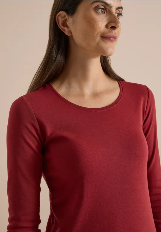 Nahaufnahme einer Frau in rotem, langärmeligem Shirt vor neutralem Hintergrund.