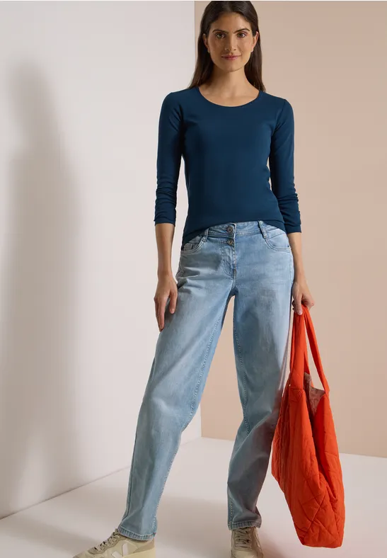Frau in dunkelblauem Langarmshirt und hellen Jeans, hält eine rote Einkaufstasche.