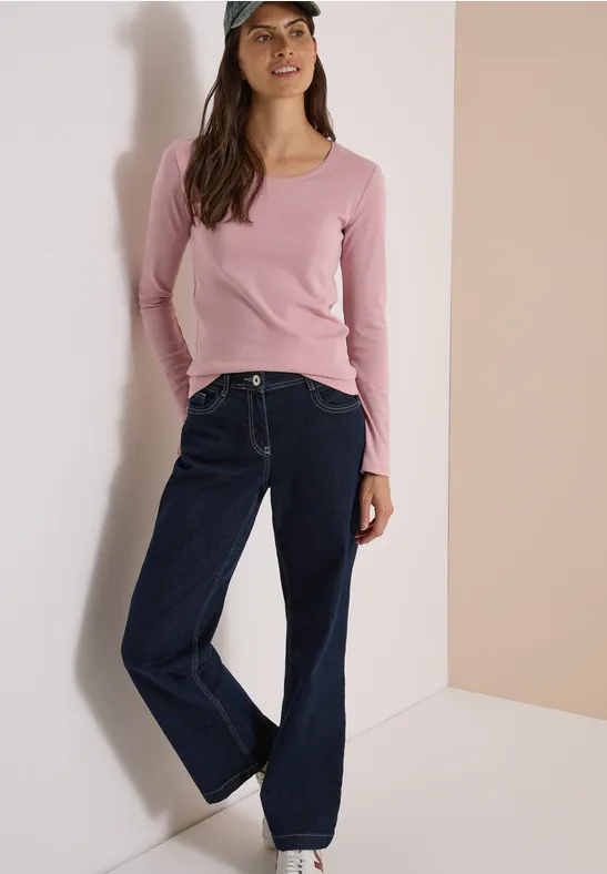 Eine Frau mit rosafarbenem Langarmshirt, weiten Jeans und Cap lehnt an einer Wand.
