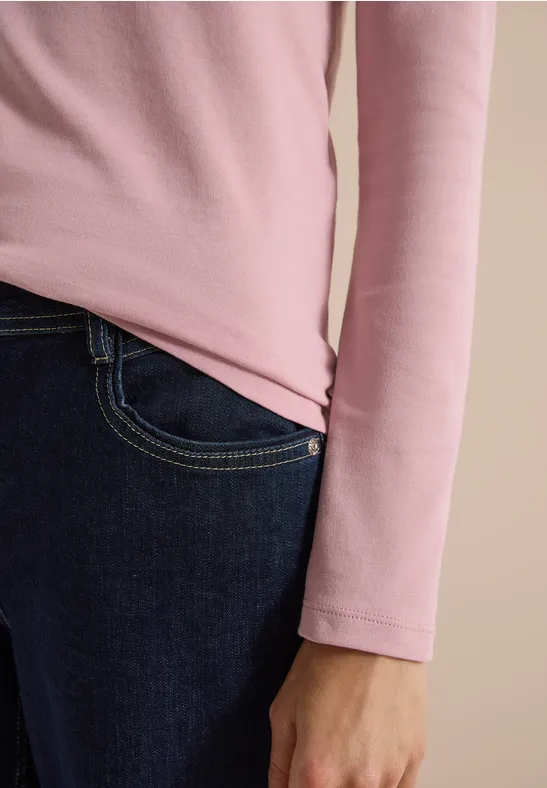 Nahaufnahme einer Person in rosa langem Oberteil und dunkler Jeans, Hand seitlich am Körper.