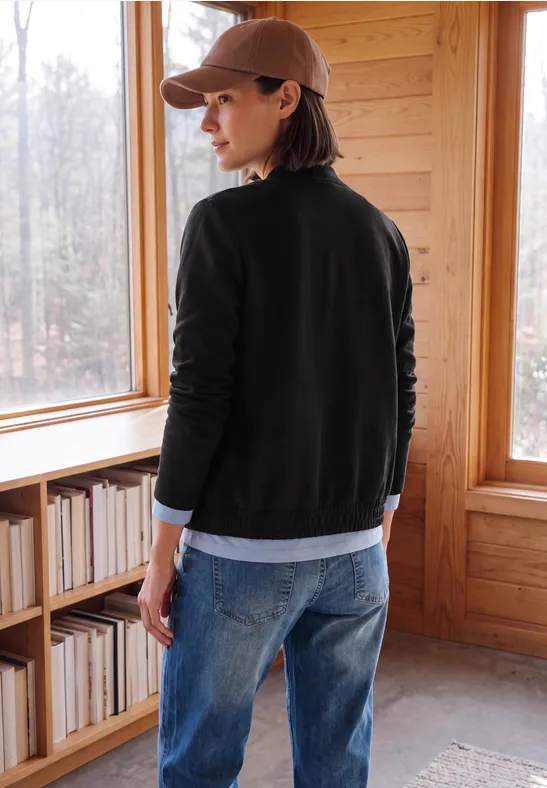Personne en casquette et veste noire, denim, dans une pièce en bois, regardant dehors par la fenêtre.