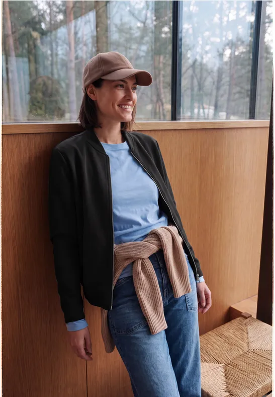 Une femme souriante en casquette, veste bleue et jean, appuyée sur une fenêtre dans un intérieur lumineux.