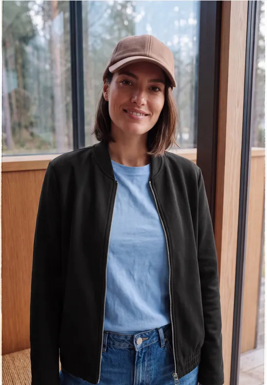 Jeune femme souriante en casquette, veste noire et jean, devant une grande fenêtre en bois.