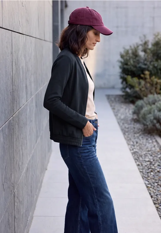Frau mit Baseballcap, schwarzer Jacke und Jeans lehnt seitlich an einer Mauer im Freien.