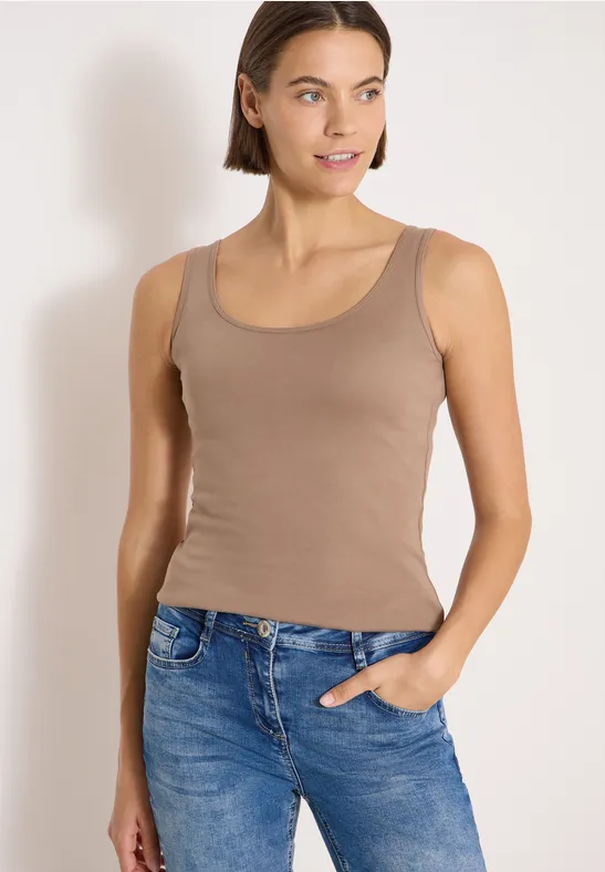 Junge Frau trägt braunes Tanktop und blaue Jeans, eine Hand in der Tasche, vor hellem Hintergrund