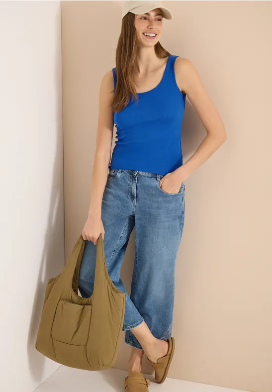 Une femme porte un débardeur bleu, un jean ample et tient un sac à main vert olive.