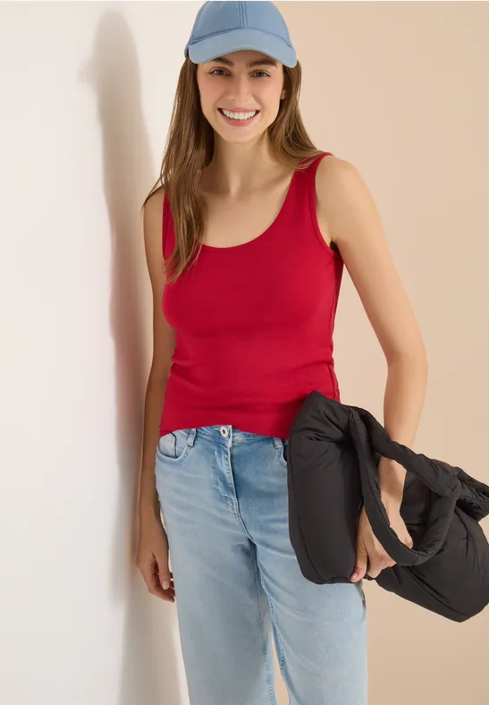 Femme en débardeur rouge et jean appuyée contre un mur, sac noir à l'épaule.
