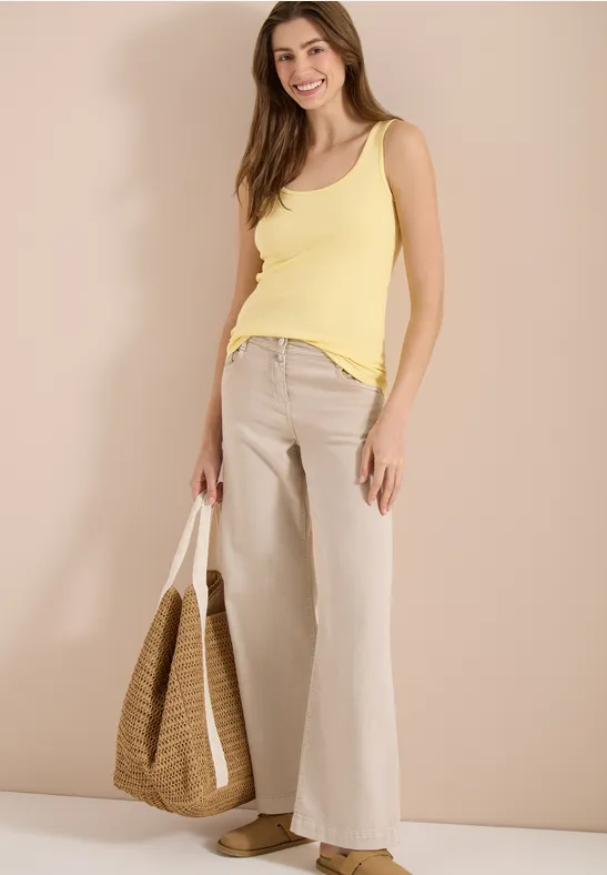 Vrouw in geel hemd en beige wijde broek met gevlochten tas, staand voor een lichte achtergrond.