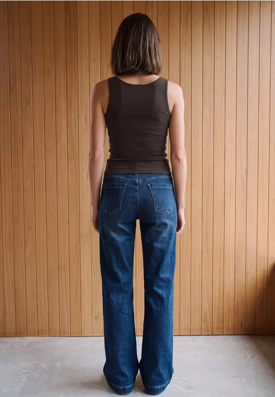 Eine Frau steht vor einer Holzvertäfelung, trägt schwarzen ärmellosen Top und blaue Jeans, Rückenansicht.