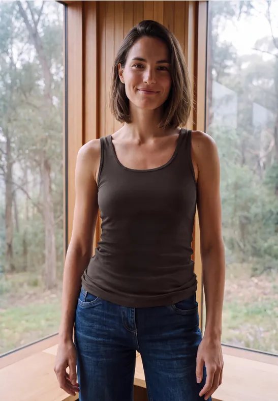 Eine lächelnde Frau in Tanktop und Jeans vor großem Fenster mit Wald im Hintergrund.