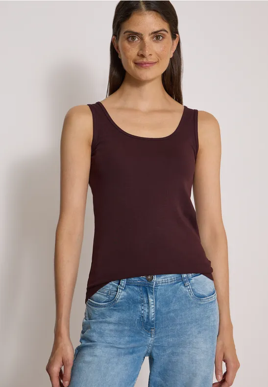 Vrouw staat voor neutrale achtergrond, draagt een bordeaux top en blauwe jeans.