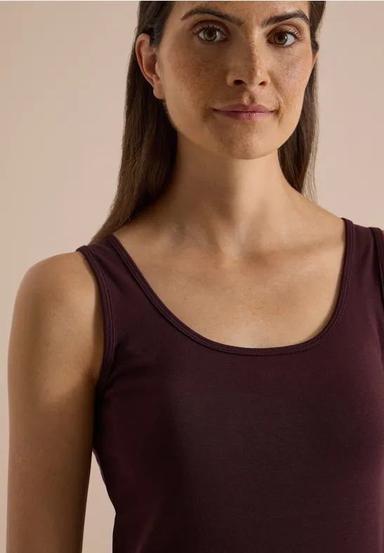 Een vrouw met bruin haar draagt een bordeaux mouwloos topje voor een lichte achtergrond.