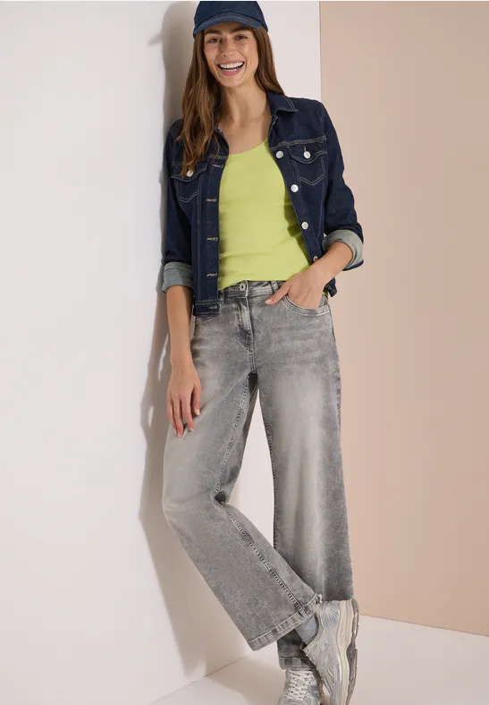 Vrouw geleund tegen muur, jeansjack, groen topje, wijde grijze jeans en witte sneakers.