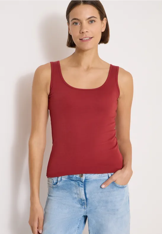 Femme en débardeur rouge et jean bleu clair devant un mur clair, une main dans la poche.