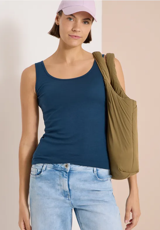 Femme en débardeur bleu et jean clair, sac olive à l'épaule, devant un fond neutre.