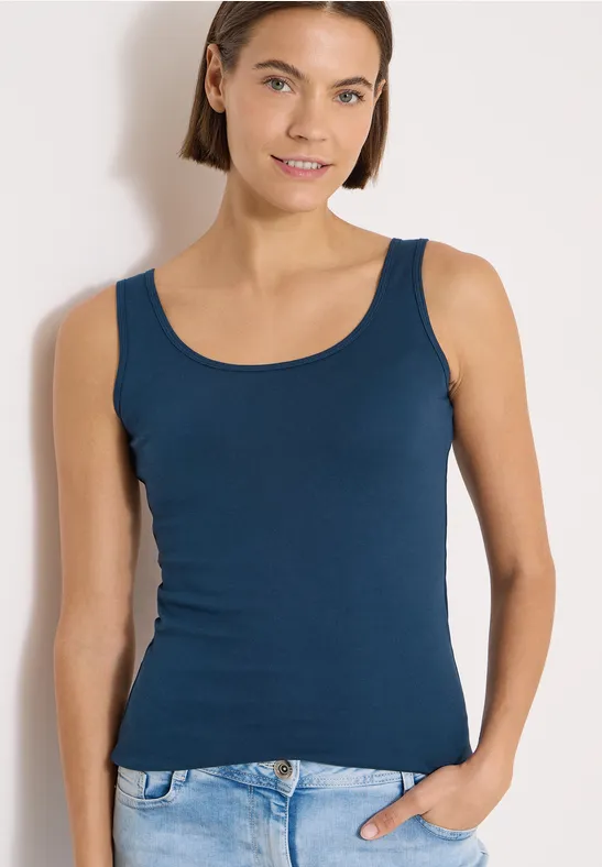 Femme portant un débardeur bleu ajusté et un jean, posant devant un fond clair.