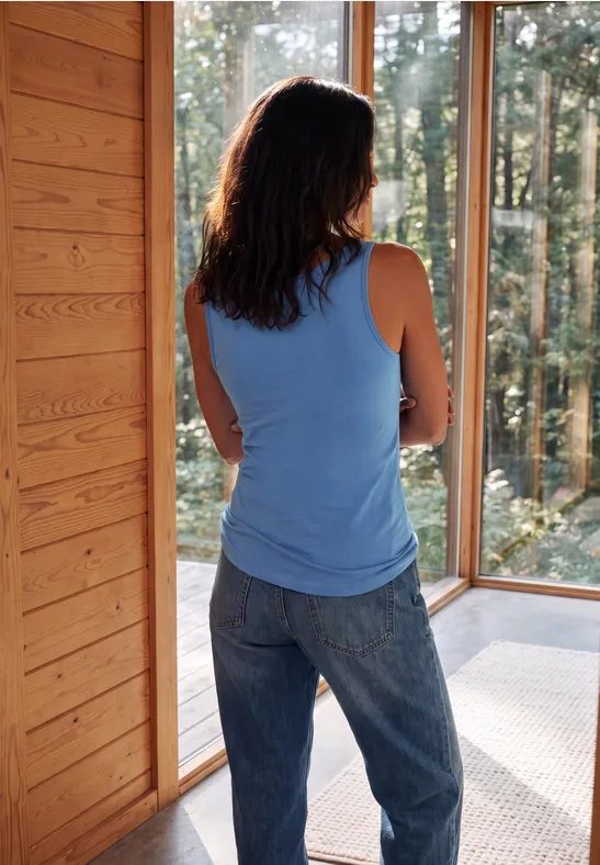 Frau in blauem Top und Jeans steht in Holzhütte und blickt durch große Fenster in den Wald.