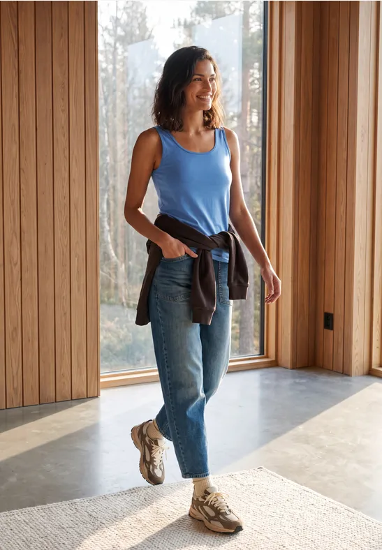 Frau in blauem Top und Jeans steht in modernem Raum mit großen Fenstern.
