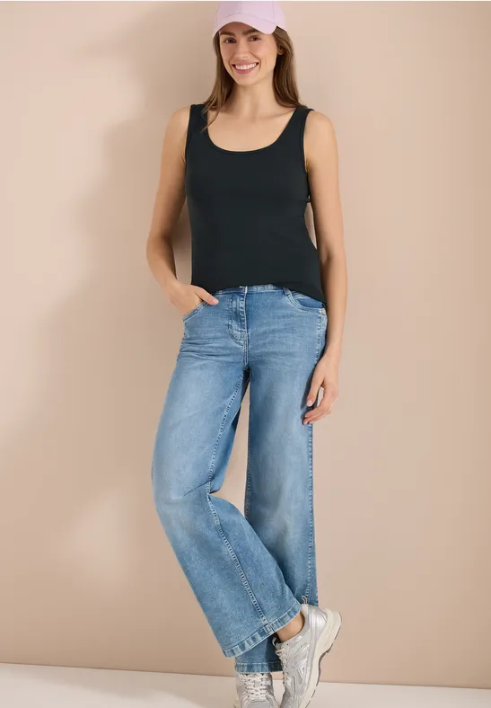 Frau trägt schwarzes Top, weite blaue Jeans und weiße Sneaker vor neutralem Hintergrund.