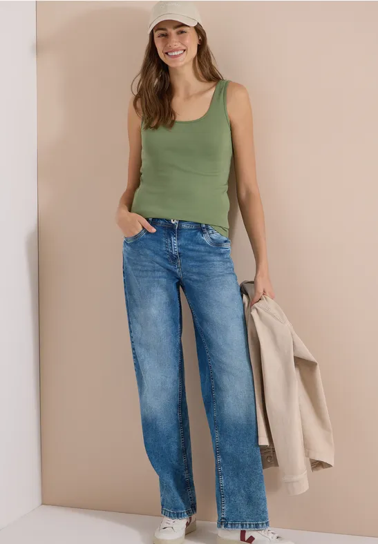 Vrouw in groen topje en blauwe jeans leunt tegen een beige muur, houdt een beige jas vast.