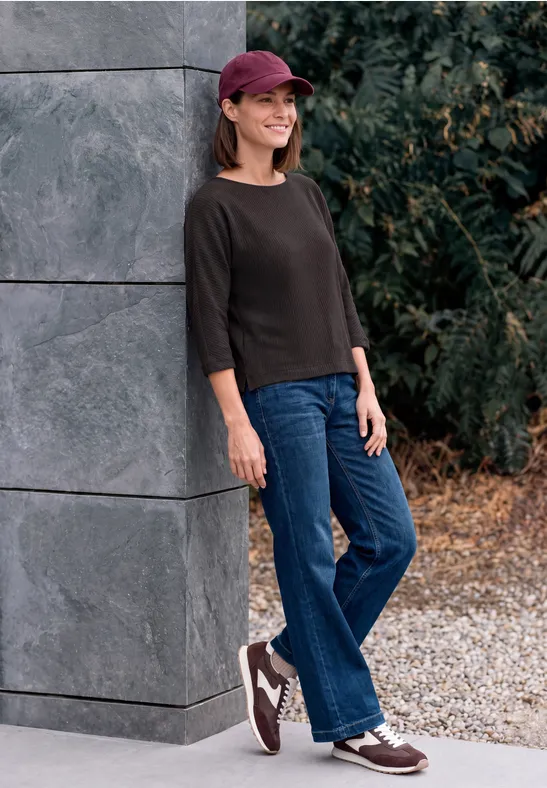 Eine Frau lehnt lächelnd an einer grauen Säule, trägt Jeans, dunkles Oberteil und eine bordeauxrote Kappe.