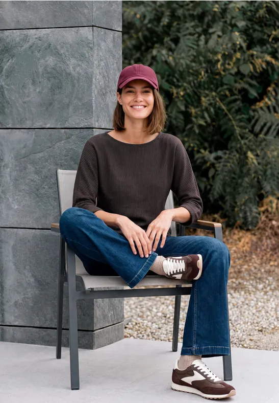 Eine lächelnde Frau sitzt auf einem Stuhl, trägt Jeans, schwarze Bluse, Sneaker und rote Mütze.