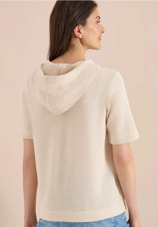 Vrouw van achteren in een korte, gebreide crèmewitte hoodie tegen een neutrale achtergrond.