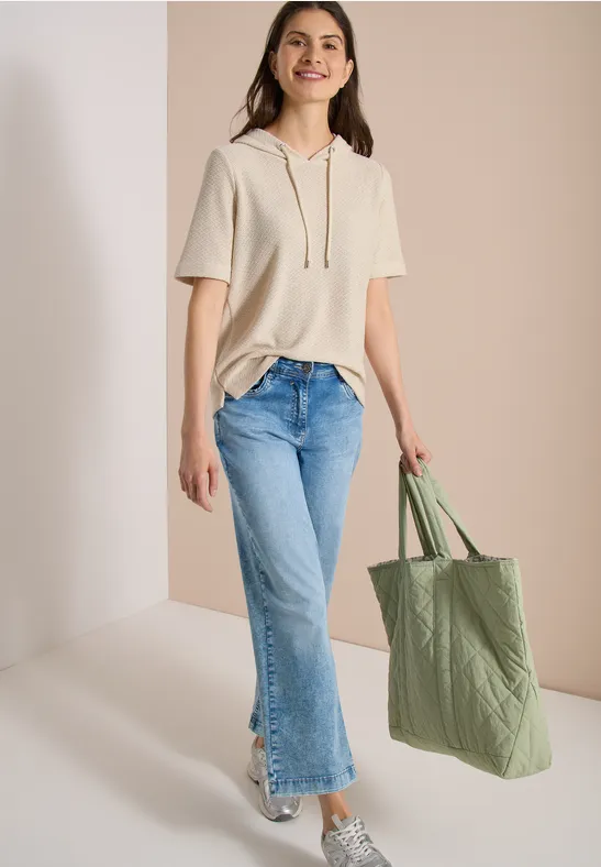 Vrouw in korte beige hoodie, lichte jeans en sneakers, met een groene stoffen tas.