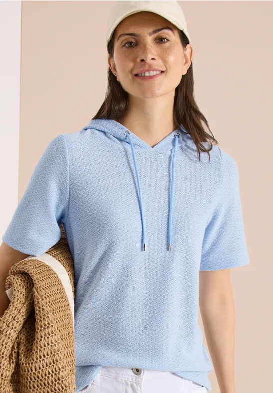 Vrouw draagt lichtblauw kort mouw capuchonshirt van fijngebreide stof, houdt een beige gehaakte tas vast.