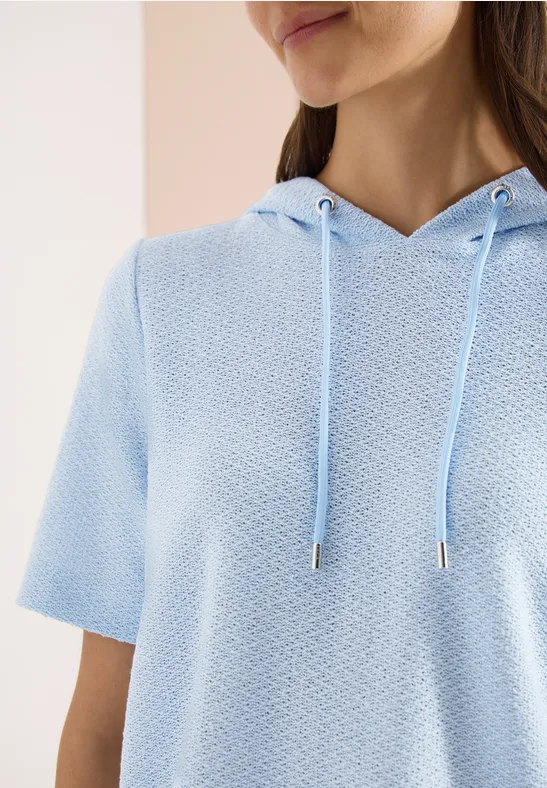 Vrouw draagt lichtblauwe, kort mouwige capuchontrui met structuur, gezicht en bovenlichaam deels zichtbaar.