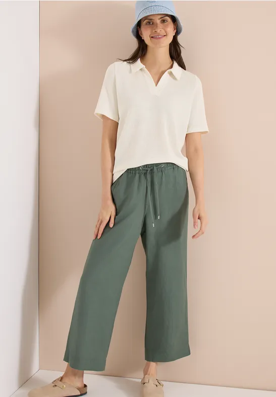 Vrouw in crème poloshirt en wijde groene stoffen broek voor effen achtergrond