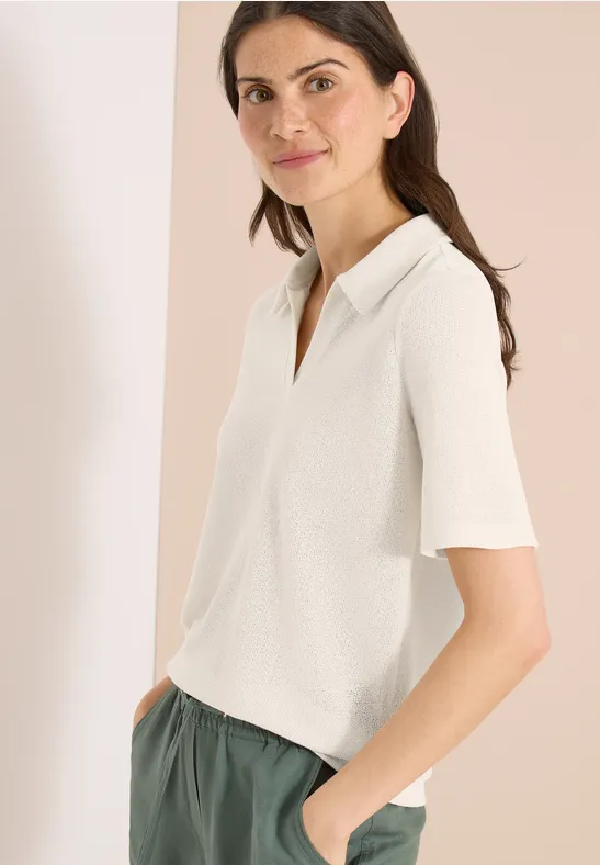 Vrouw in een witte polo met korte mouwen en een groene broek, zijwaarts staand met handen in de zakken.