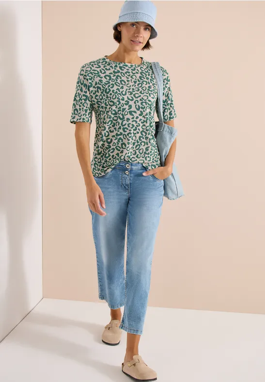 Vrouw in groen-wit gebloemde top, lichte spijkerbroek en sleehakken espadrilles, met lichtblauwe hoed en tas.