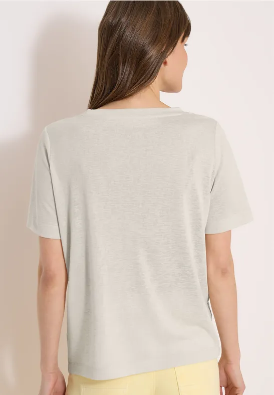 Eine Person steht mit dem Rücken zur Kamera und trägt ein beiges T-Shirt sowie gelbe Hosen vor hellem Hintergrund.