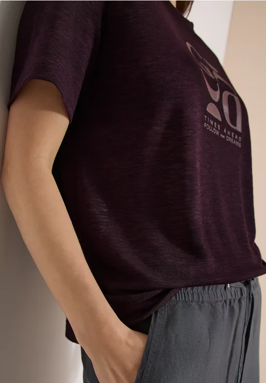 Eine lässige Pose: Person lehnt an der Wand, trägt violettes T-Shirt und graue Hose.