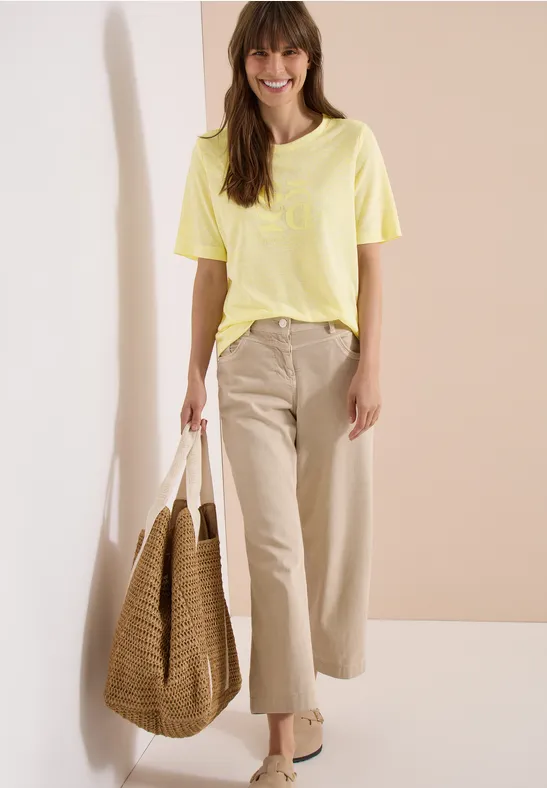 Frau in gelbem T-Shirt, beiger Hose mit großer Korbtasche vor neutralem Hintergrund.