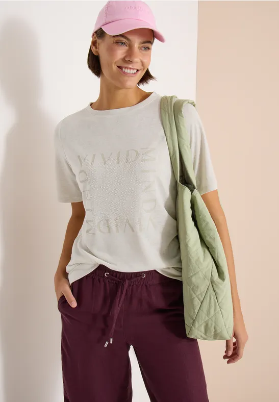 Vrouw in wit T-shirt, bordeaux joggingbroek, roze pet en groene gewatteerde tas.